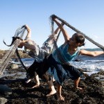 three dancers tangled intentionally in fishing net bend over at the waist with their backs to each other on the rocks in Maine.