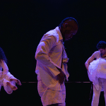 Five dance in white costumes perform on a dimly lit stage, illuminated by blue and purple theatrical lighting.