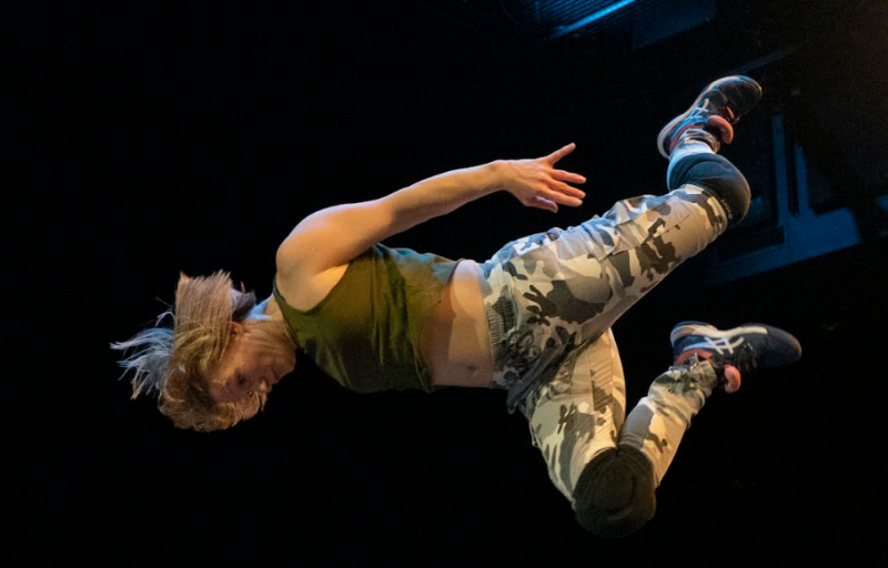 a dancer wearing a green top and camo pants jumps in a horizontal energetic position with arms back and knees bent