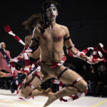a white masc presenting dancer dressed in shreds of the American Flag jumps with both knees up and to the sides with arms down