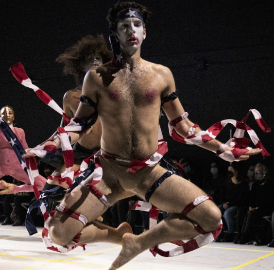 a white masc presenting dancer dressed in shreds of the American Flag jumps with both knees up and to the sides with arms down