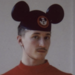 a white masc person with a moustache wears a mickey mouse hat with a red shirt and glances at the camera with a straight face 