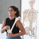 The photo shows Allegra with a skeleton model behind her. She has the right leg of the skeleton over her shoulder. 