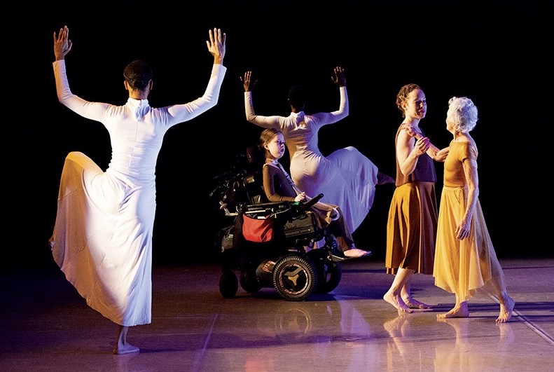 Two female dancers in long white dresses stand still with arms bent and up while a girl in a wheelchair & two women pass through