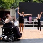 3 female identifying dancers wearing black, dancer to left using a wheelchair. 