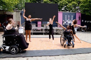 3 female identifying dancers wearing black, dancer to left using a wheelchair. 