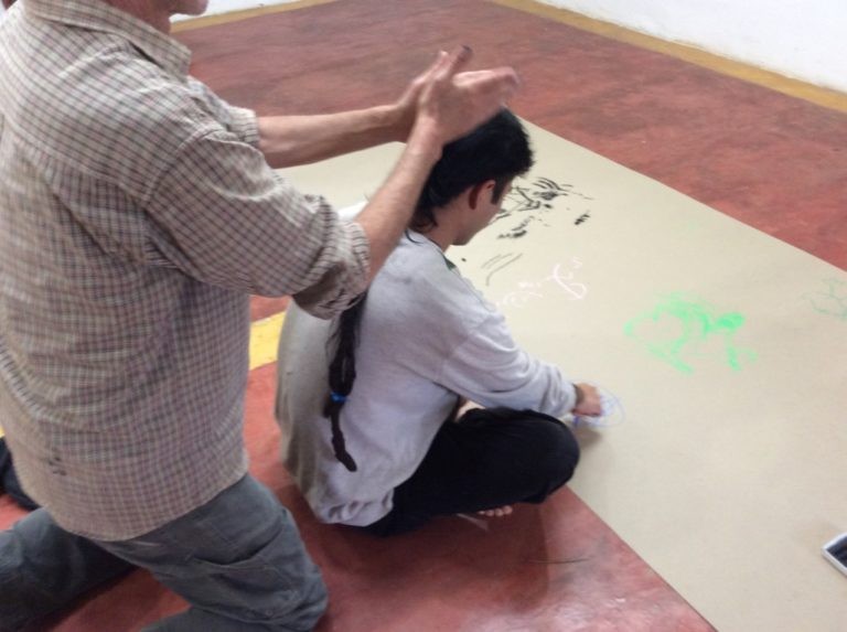 Image of a person sitting with legs crossed looking at a drawing placed on the floor with another person standing behind them.