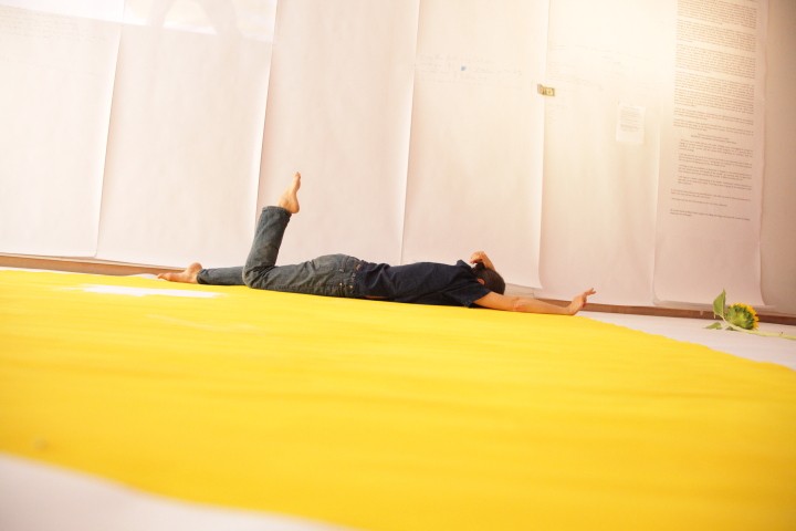 Daria Faïn lies on her stomach on a bright yellow floor. One leg is bent at a 90 degree angle with her foot pointed.