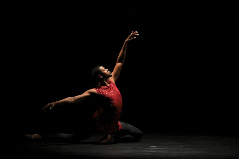 Male dancer in burgundy top and black tights sits in 4th position, back leg extended, facing stage left reaching in 4th.