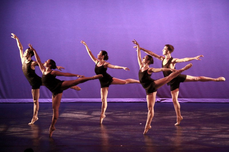 Five dancers space out on stage, facing stage right, in relevé arabesque, wearing black costumes. Stage lighting is purple.. 