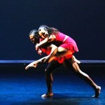 Two dancers in vibrant red costumes, female dancer is in a passionate embrace on male dancer's back with leg extended forward.
