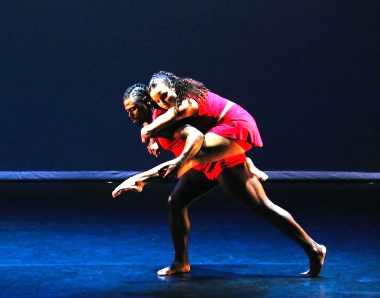Two dancers in vibrant red costumes, female dancer is in a passionate embrace on male dancer's back with leg extended forward.