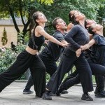 Dancers dressed in black, holding each other in a V formation, faces upward towards the sky