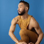 An Asian Male dancer wearing yellow with a blue background posing on a stool.