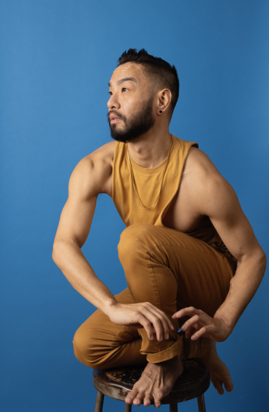 An Asian Male dancer wearing yellow with a blue background posing on a stool.