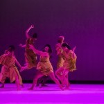 Seven dancers on stage dancing in green costumes under purple lighting