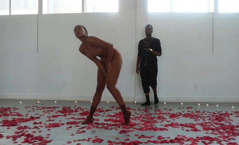 Women dressed in nude clothing stands in the middle of the room with rose petals sprawled across the floor.