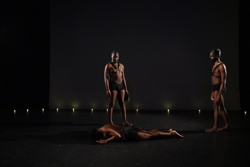 Two dancers stand starkly light over a dancer laid prone on the floor
