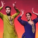 The image shows two South-Asian dancers, one male and one female, performing on stage, wearing traditional Indian attire.