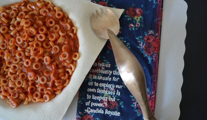 A bowl of Spaghetti-Os next to an embroidered tablecloth with a Candida Royalle quote