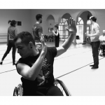 A dancer in a wheelchair looking down over his shoulder with arms uplifted towards his other shoulder.