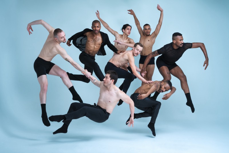 7 dancers in black rehearsal attire jump, with interconnecting limbs