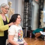 Ann Rodiger, standing, places her hands along the neck of a seated student during a workshop at the Balance Arts Center