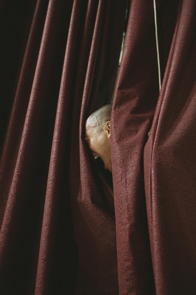 between the folds of a curtain a face in profile screams into the dark red fabric. The person is bald and Asian.