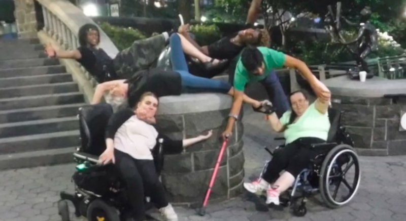 Two white women in wheelchairs leaning toward a stone wall. Two black men, an Asian man and white elder woman draped atop it.