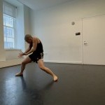 A dancer in a white-walled dance studio pressing downward into the floor from a standing squat position.