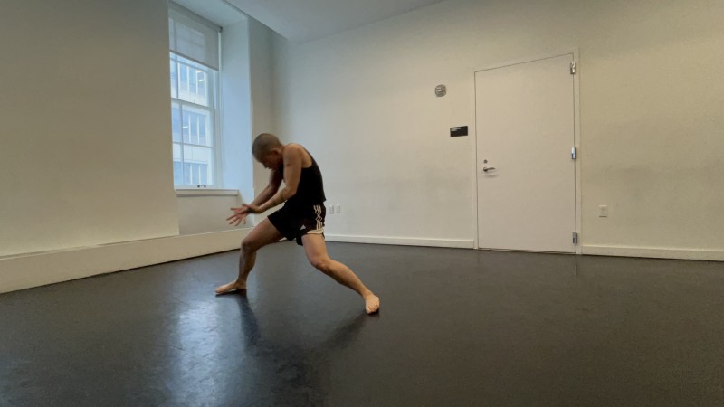 A dancer in a white-walled dance studio pressing downward into the floor from a standing squat position.