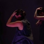 Three dancers stand facing the side in shadowed, pink light, one hand on their head and the other on their heart.