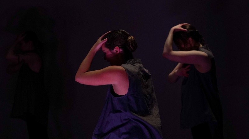 Three dancers stand facing the side in shadowed, pink light, one hand on their head and the other on their heart.