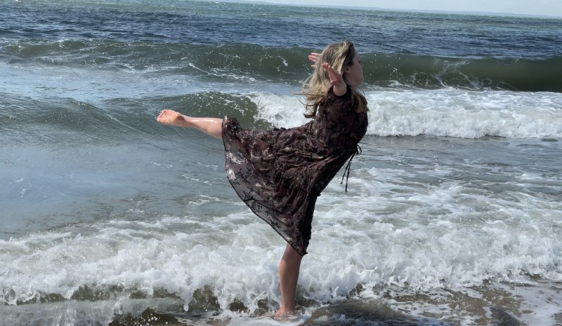 A dancer wearing a flowing, brown dress splashes in the ocean.