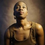 Black woman with short hair in brown dress poses in front of brown back drop looking up in a portrait photo.