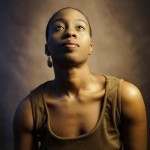 Black woman with short hair in brown dress poses in front of brown back drop looking up in a portrait photo.