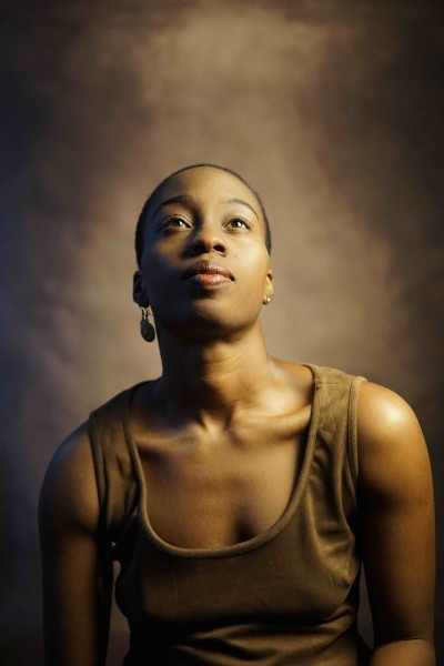 Black woman with short hair in brown dress poses in front of brown back drop looking up in a portrait photo.