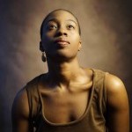 Black woman with short hair in brown dress poses in front of brown back drop looking up in a portrait photo.