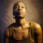 Black woman with short hair in brown dress poses in front of brown back drop looking up in a portrait photo.