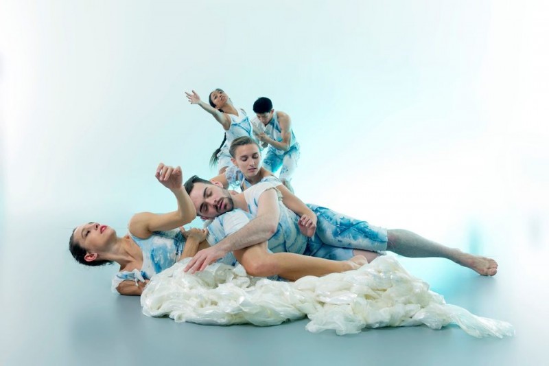 Dancers tumbling to the floor in a coiled group, wearing blues and whites, eyes closed, immersed in the sensation of a wave