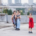 Eight dancers in a line on a rooftop, each hugging the person in front of them, with the first dancer in red and second in blue