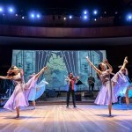 A group of dancers with violet sheer dresses and one leg extended with orchestra and violinist in the backdrop