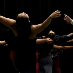 From behind, an image of dancers in a blackbox studio hinged and arched toward the sky with arms overhead. 