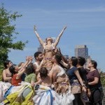 An image of a group of dancers wearing costumes made from recycled plastics, hold up a singular dancer in the air.