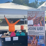 Artistic Director Kristina Bermudez is wearing a neon orange shirt in front of a sign that says join us. Outdoor table event.