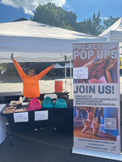 Artistic Director Kristina Bermudez is wearing a neon orange shirt in front of a sign that says join us. Outdoor table event.