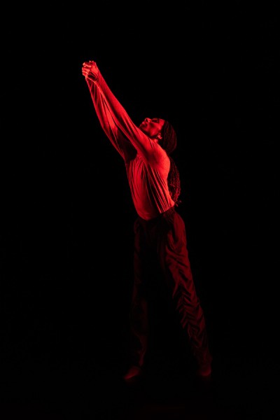 Ke'ron Wilson shining through red lighting on a dark stage, hands clasp and reaching up and out.