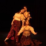 Three dancers dressed in white and maroon create a family portrait like tableaux with their chest and gaze forward.