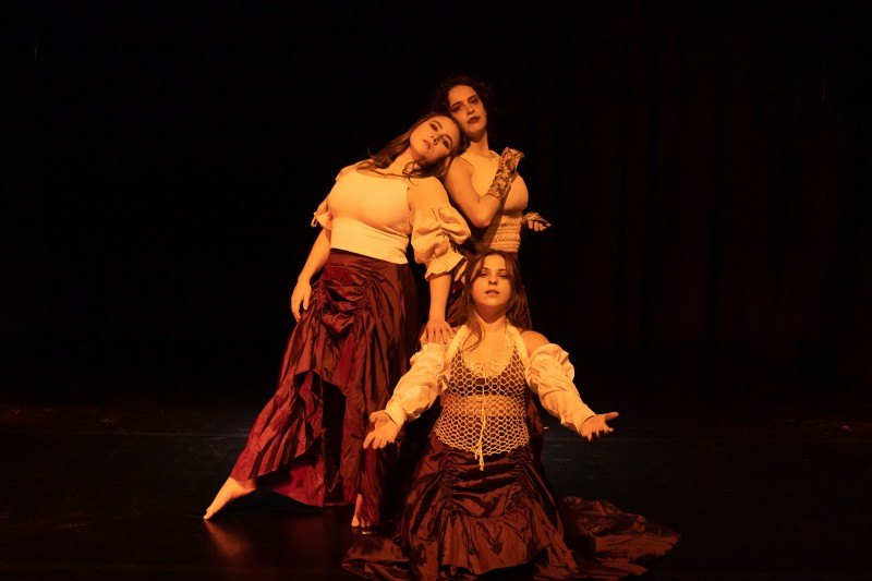 Three dancers dressed in white and maroon create a family portrait like tableaux with their chest and gaze forward.