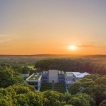 Bird's eye view of The Watermill Center, a large building surrounded by woods in front of an orange sunset. 